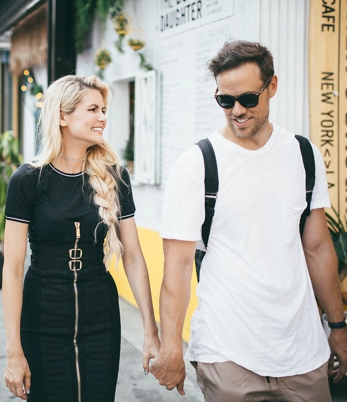 A happy couple walks hand in hand in an urban setting, showcasing a casual and stylish look. The woman wears a fitted black dress with a zipper detail, while the man sports a white t-shirt and beige shorts, complemented by sunglasses and a backpack. The background features a vibrant wall with greenery and artistic elements, enhancing the lively atmosphere. This image captures a moment of joy and connection, perfect for lifestyle and fashion content.