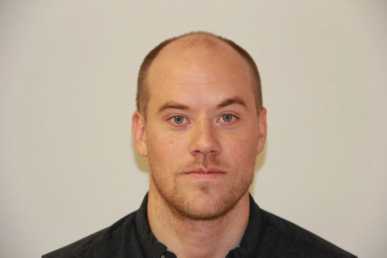 Image of a man with short, light brown hair and a slight beard, looking directly at the camera. He is wearing a black collared shirt and has a neutral expression. The background is plain and light-colored, providing a clear focus on his face. This image can be used for professional profiles or team pages, highlighting personal attributes in a corporate setting.
